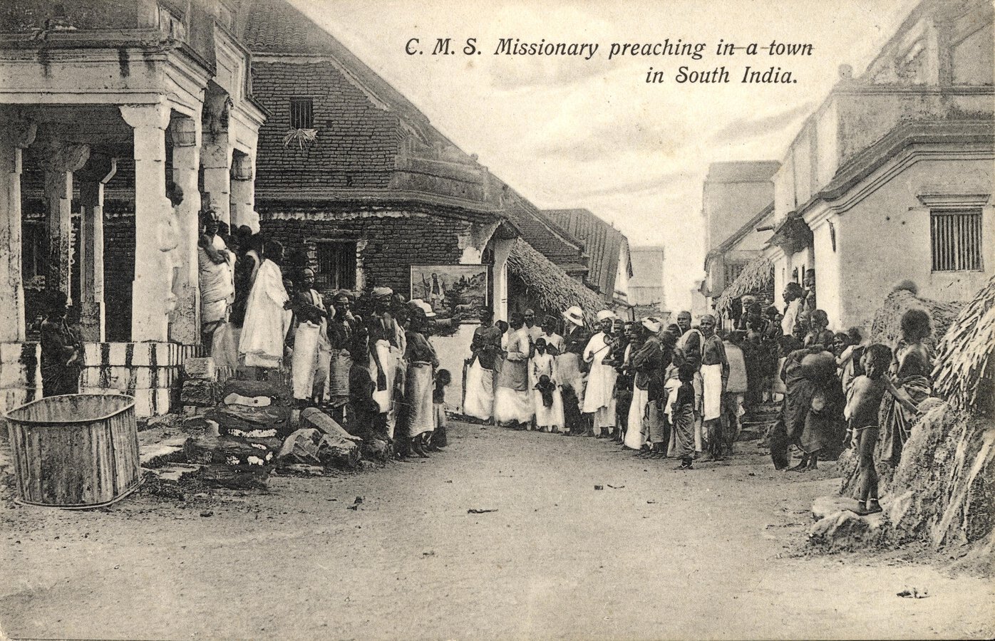 Group of people standing across the width of a street listening to a missionary who is not visible.