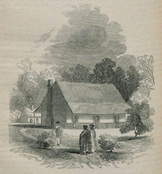 English-style house with three individuals in front and trees and bushes at sides.