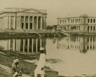 Two men, one squatting, one standing, before a small pond behind which are Greek-style buildings.