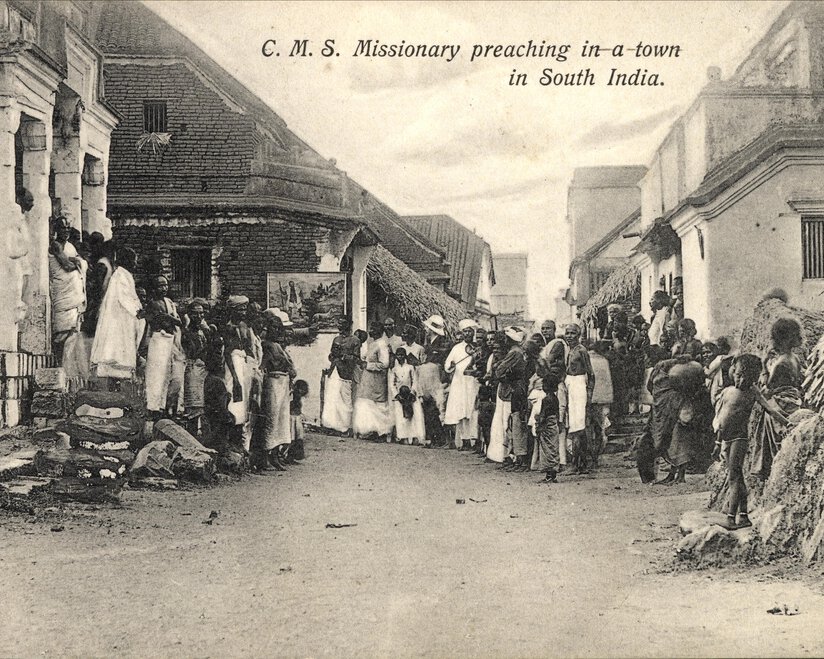 Group of people standing across the width of a street listening to a missionary who is not visible.