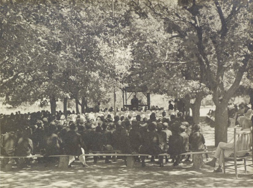 Large group of people seated on benchs under trees, facing an individual on raised stand with roof.