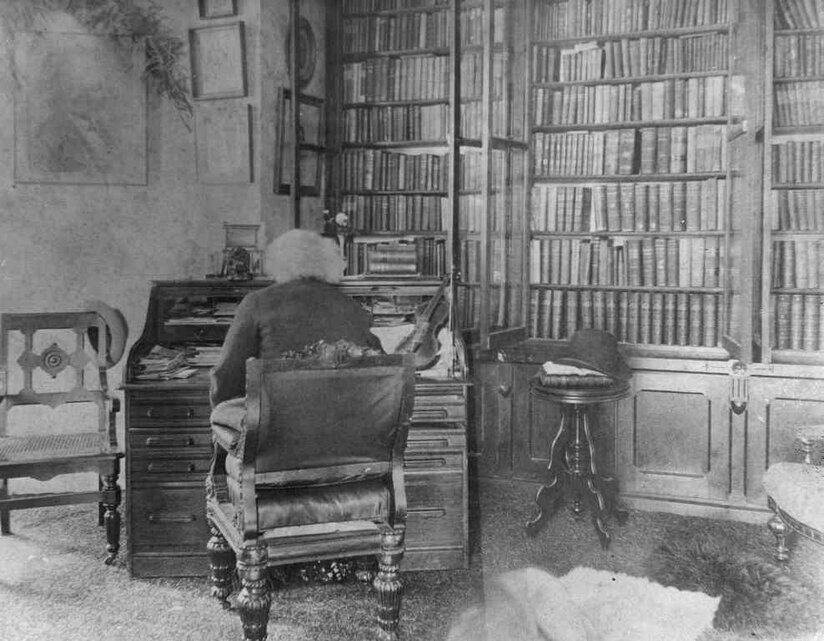 Frederick Douglass with his back to the viewer at his desk in his library with books in background.
