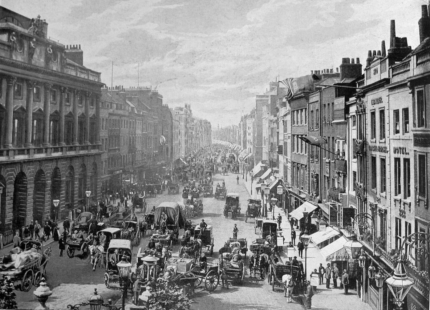 Bird's eye view of street, buildings on both sides, people and horse-drawn carriages in the street.