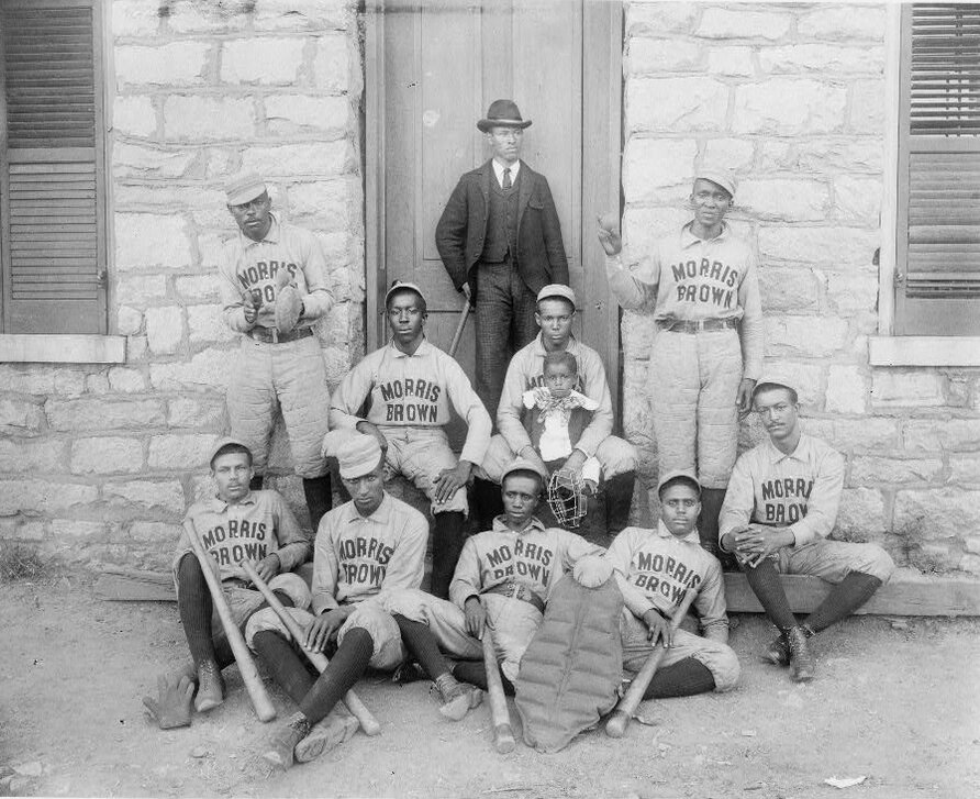 Group of seated and standing men in baseball uniforms in front of a stone building with a child and a man in a suit.