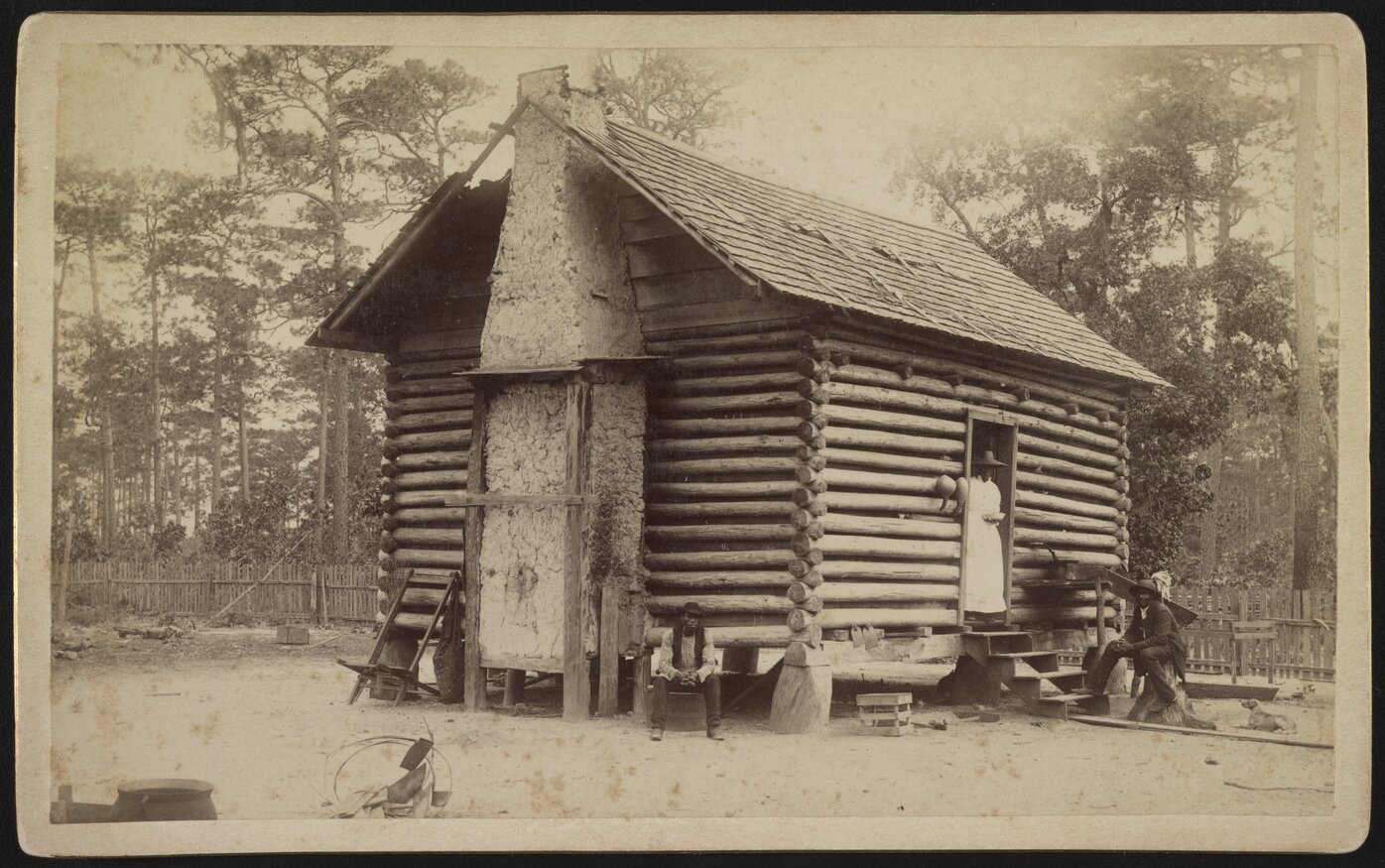 Two men sitting outside a log cabin with a woman standing in the doorway.