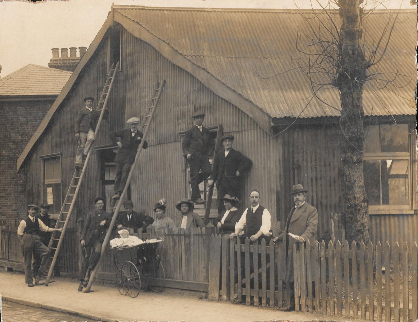 Group of people standing in front of a house with a fence, with men on ladders and a baby in a pram.