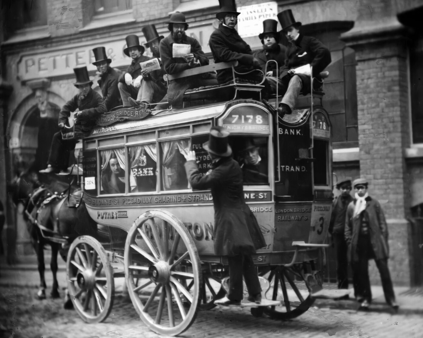 Men atop a stage coach omnibus, with horses and a driver standing in back.