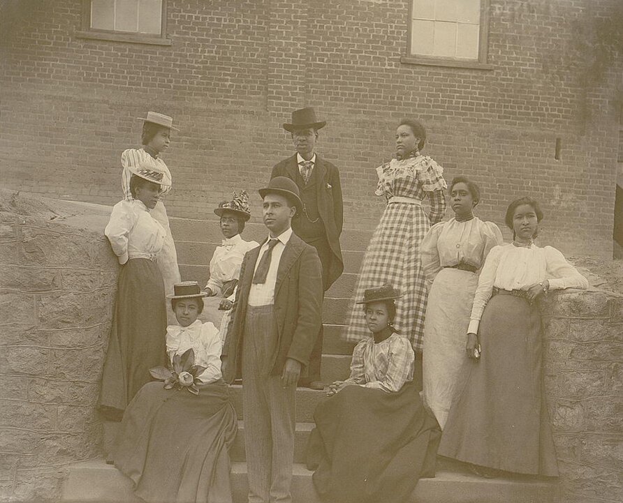 Group of men and women sit and stand on a small staircase in front of brick building.