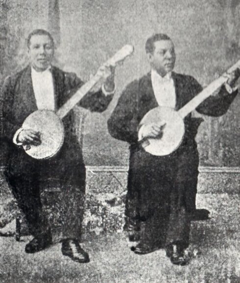 Two men in suits seated and playing banjos.