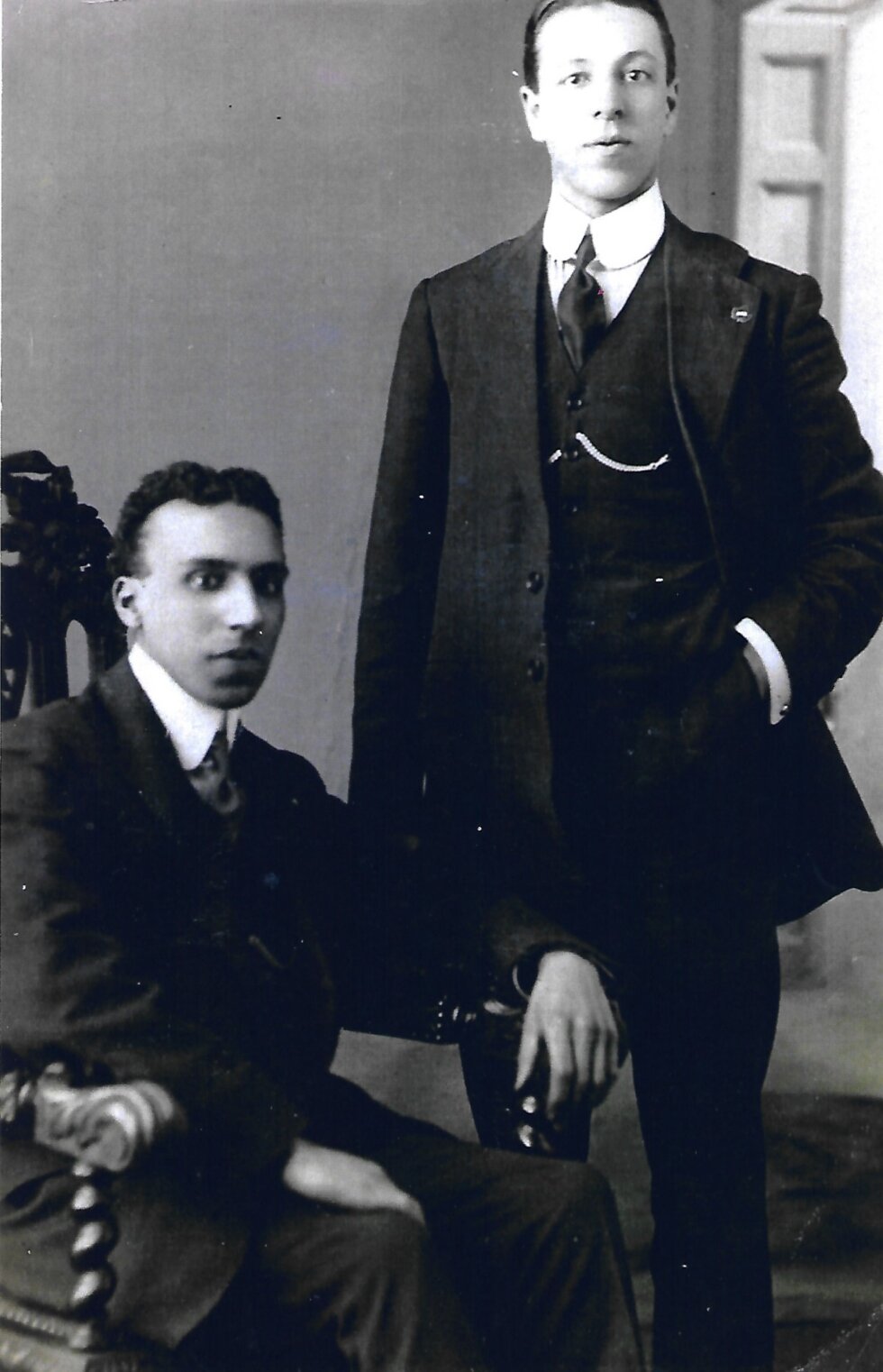 Two men in suits; one is standing and one Is seated.
