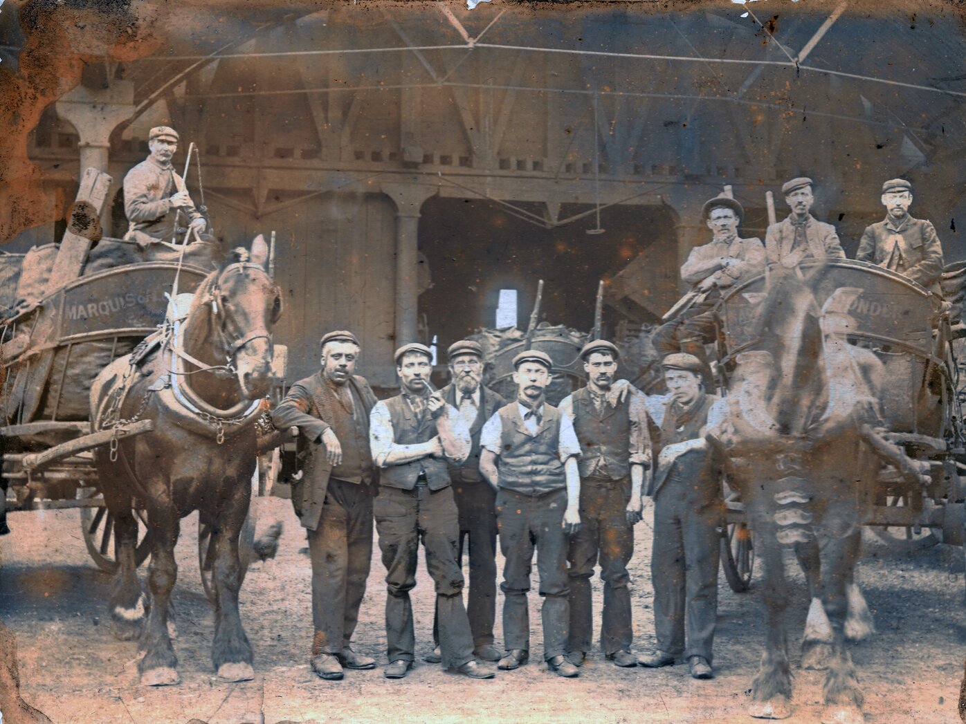 Six men standing in front of a wooden building flanked by two horse carts with men in them.