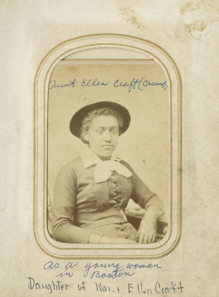 Portrait of a seated woman wearing a hat, neck scarf, and dress, looking at the camera.