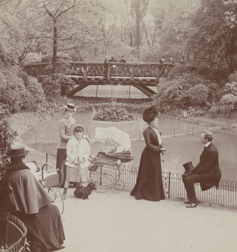 Group of men, women, children, and a pram in garden path next to small water way with a bridge in the background.