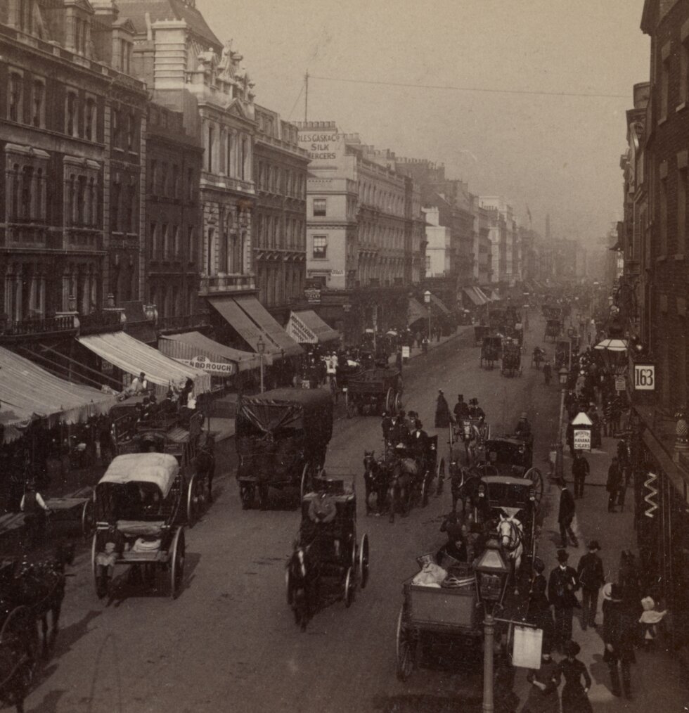 Bird's eye view of street, buildings on both sides, people and horse-drawn carriages in the street.