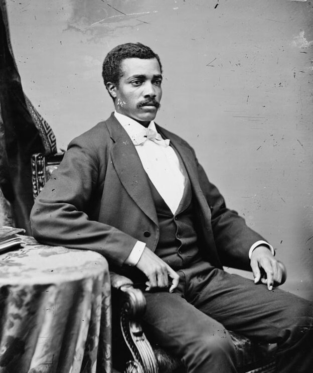 Portrait of a man in a suit seated with one arm on a table looking away from the camera.