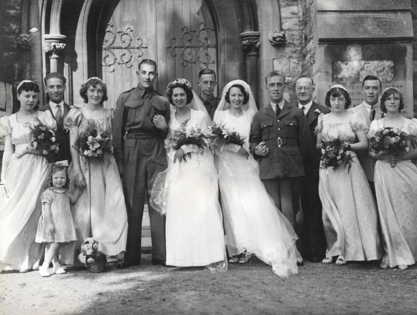 Group of people dressed in wedding attire and fancy clothes standing in front of a stone church facade.