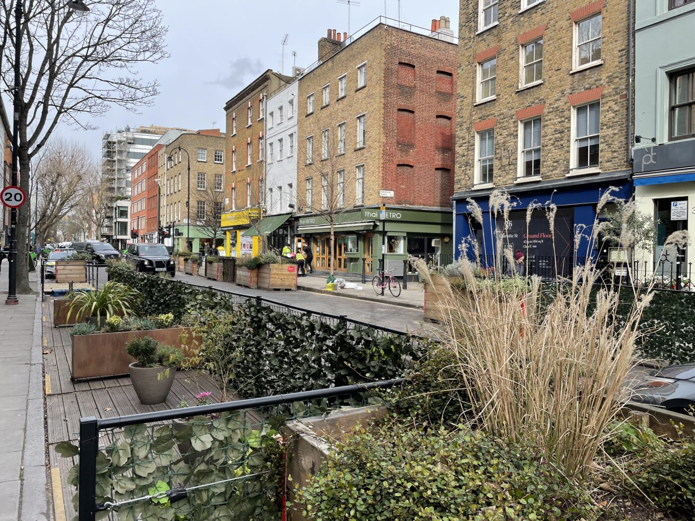 View down a city street with brick buildings on one side and plants on the other.