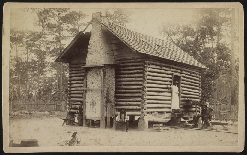 Two men sitting outside a log cabin with a woman standing in the doorway.