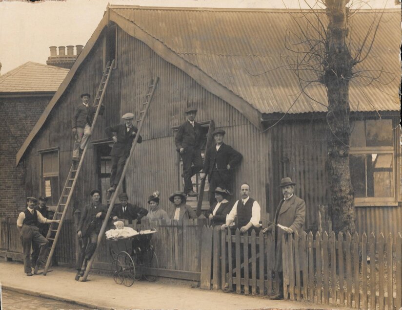 Group of people standing in front of a house with a fence, with men on ladders and a baby in a pram.