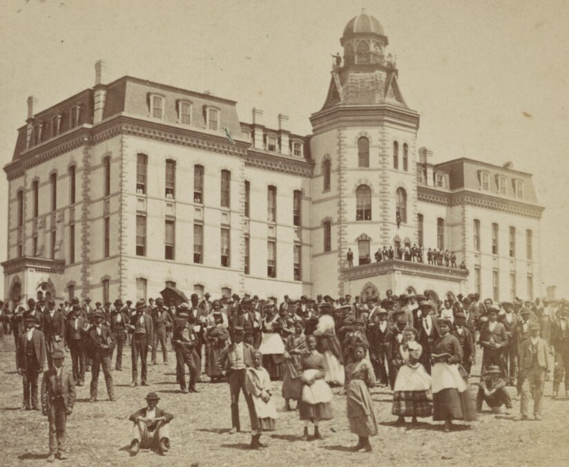 Group of men and women standing on a lawn in front of large stone building.