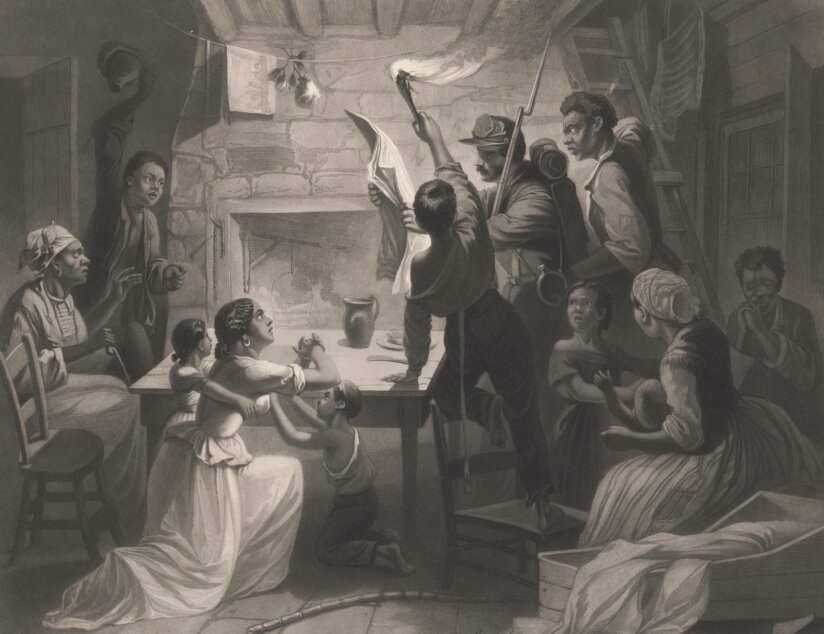 A group of people gather around a table as a soldier reads a large piece of paper by torchlight.