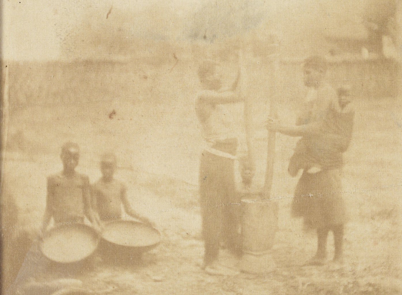 Individuals in African village. Two hold large plates; the others surround domestic implements.