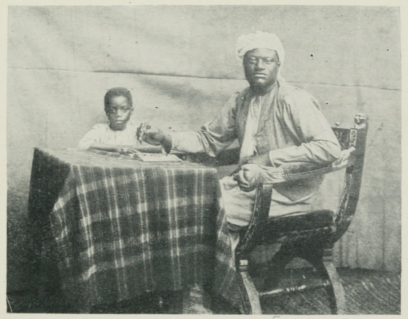 Apolo Kagwa and his son, both seated at a table; Kagwa holds a pen above a notebook.