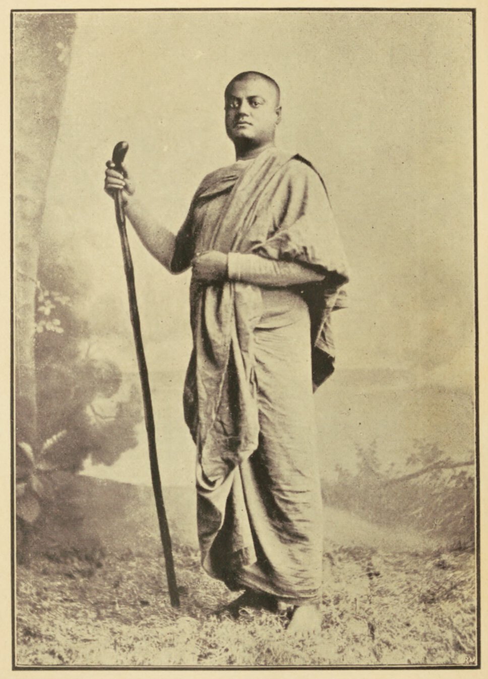 Full-body portrait of Swami Vivekanada, standing, his body turned right, holding a walking stick.