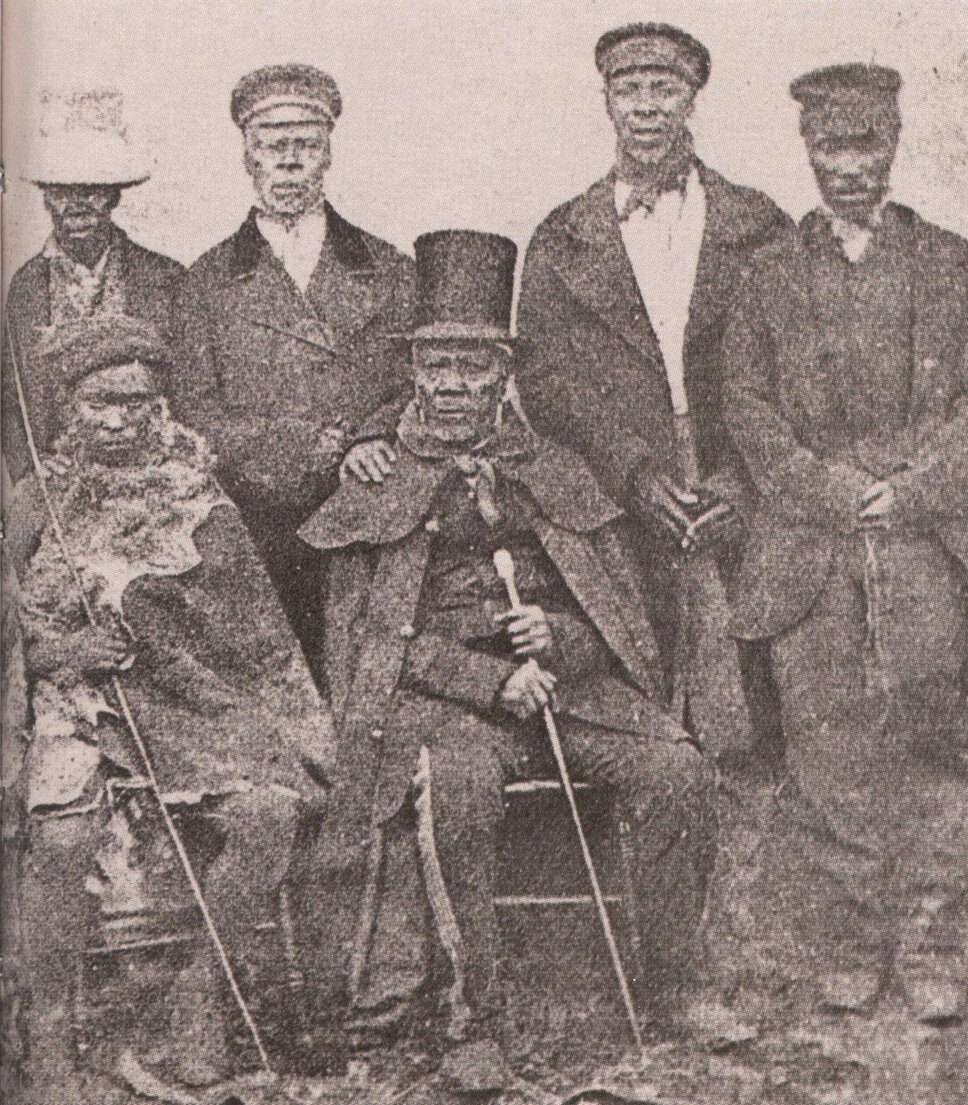 Moshoeshoe I seat and in formal attire, surrounded by five individuals, some seated, some standing.