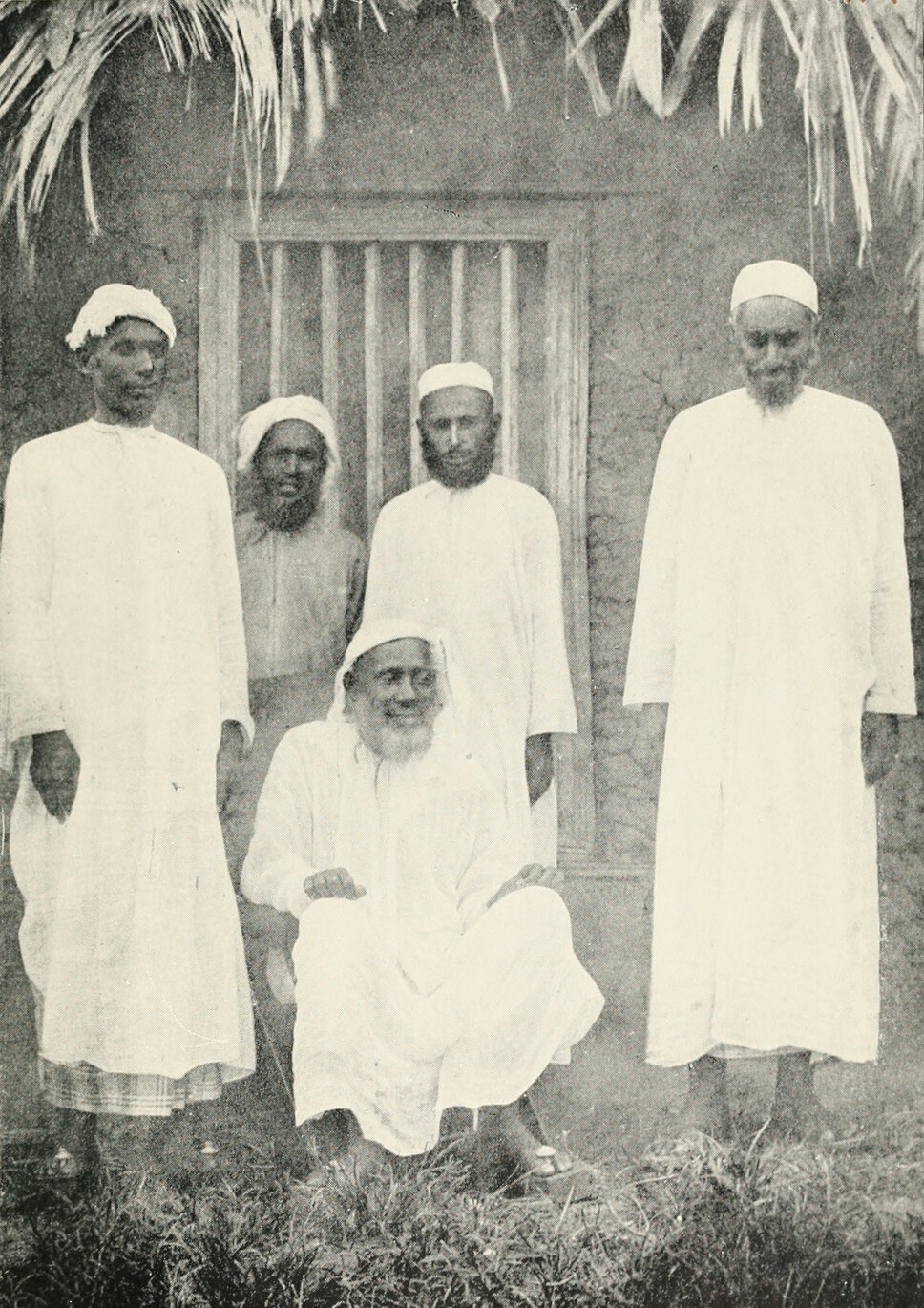 Tippu Tip seated and surrounded by four individuals, all of whom are standing.