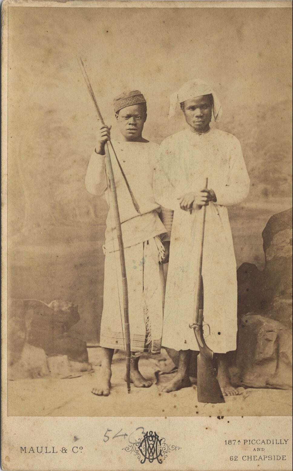 James Chuma and Abdulla Susi, in traditional clothing, with weapons, against a backdrop of rocks.