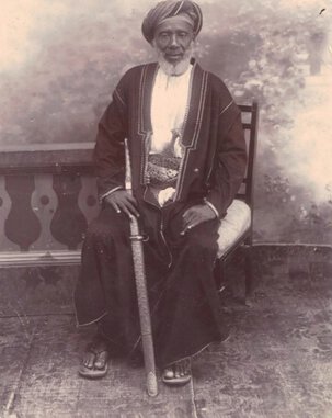 Full-body portrait of Tippu Tip, seated, facing forward, and holding a long, sheathed sword.