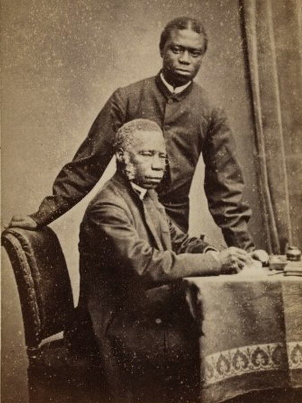 Samuel Ajayi Crowther at desk in half profile; Dandeson Crowther standing behind and facing forward.