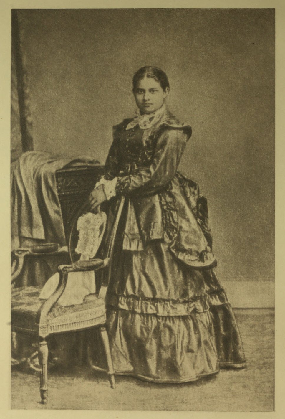 Toru Dutt standing in three-quarter profile to her right with hands on chair, but facing the viewer.