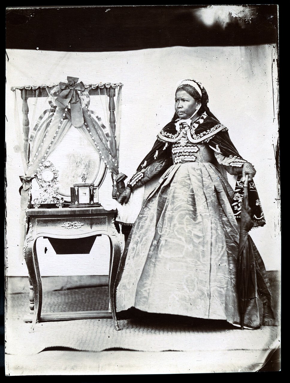Ranavalona II in regal attire, standing, looking right, with an umbrella, beside a table and mirror.