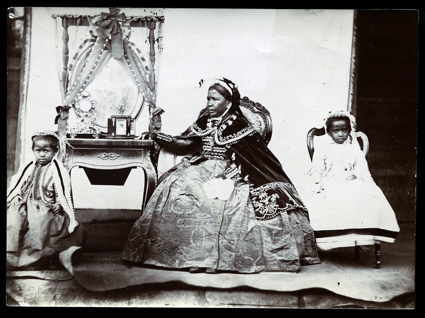 Ranavalona II in regal attire, seated, looking right, beside a table and mirror with two children.