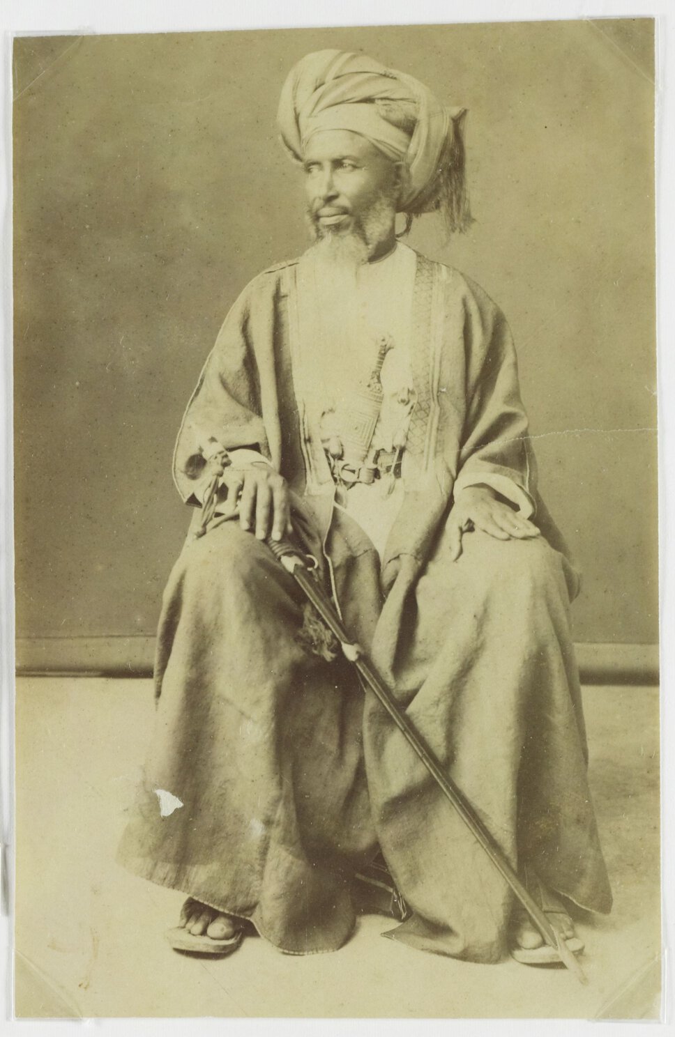 Tippu Tip, seated, looking to the right, holding a thin long sword.