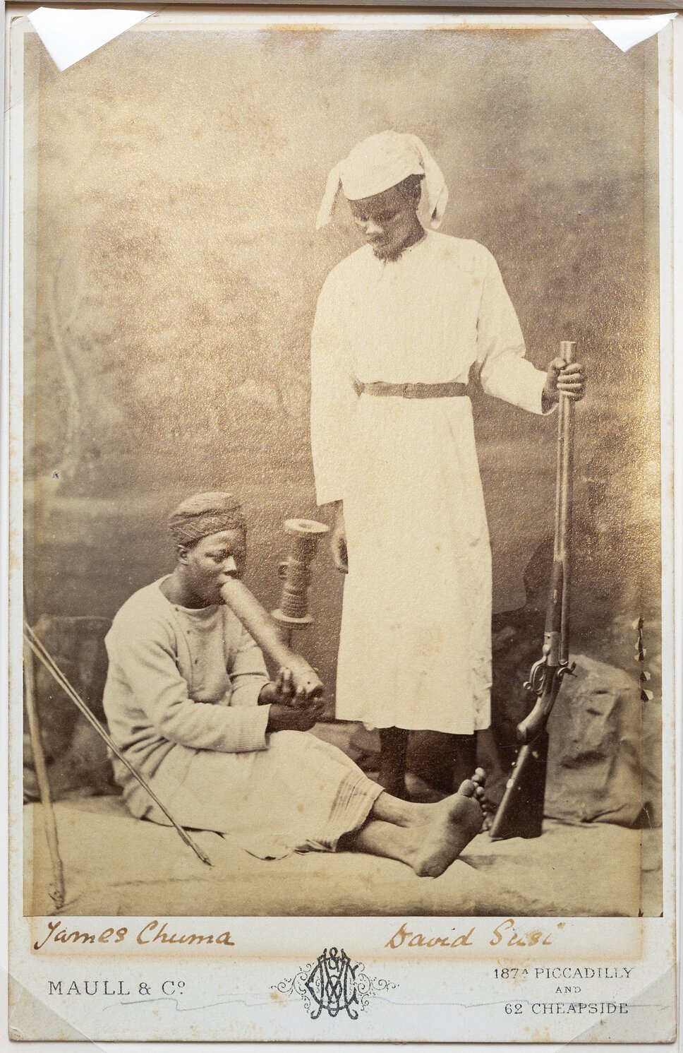 Chuma, seated with legs extended, smoking pipe and being watched by Susi who holds rifle upright.