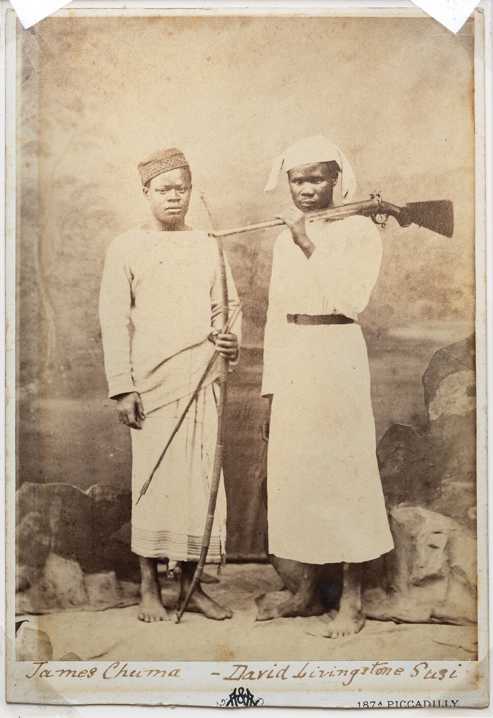 Chuma, standing, with long bow and arrow, beside Susi, with rifle across shoulder.