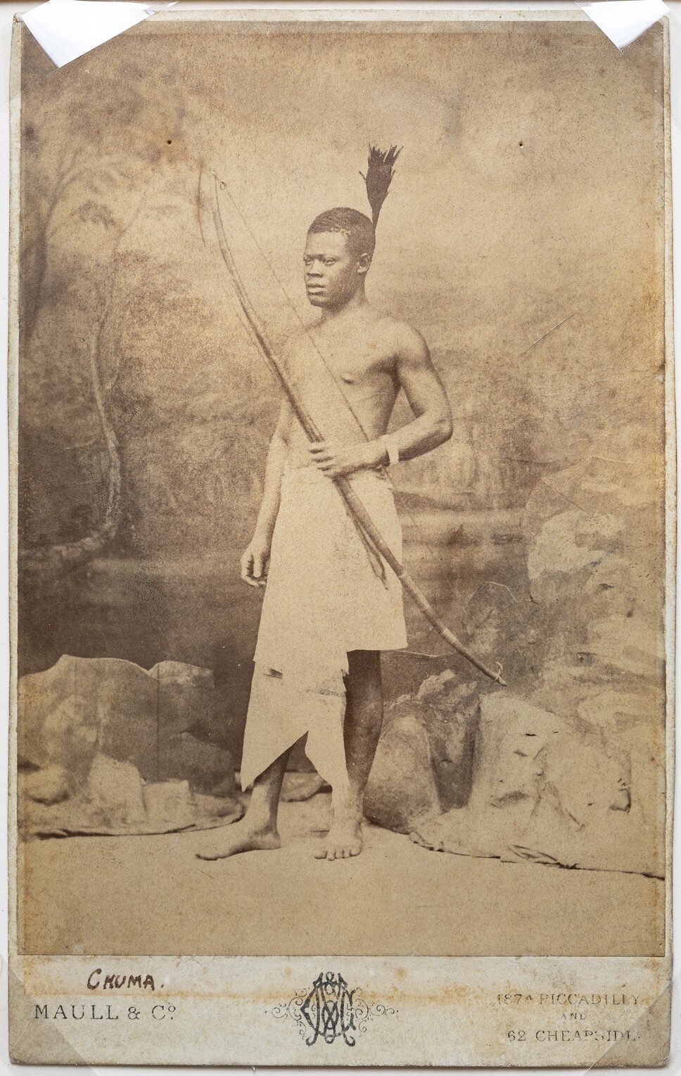 Chuma, standing, in traditional waist covering, with long bow and arrow.