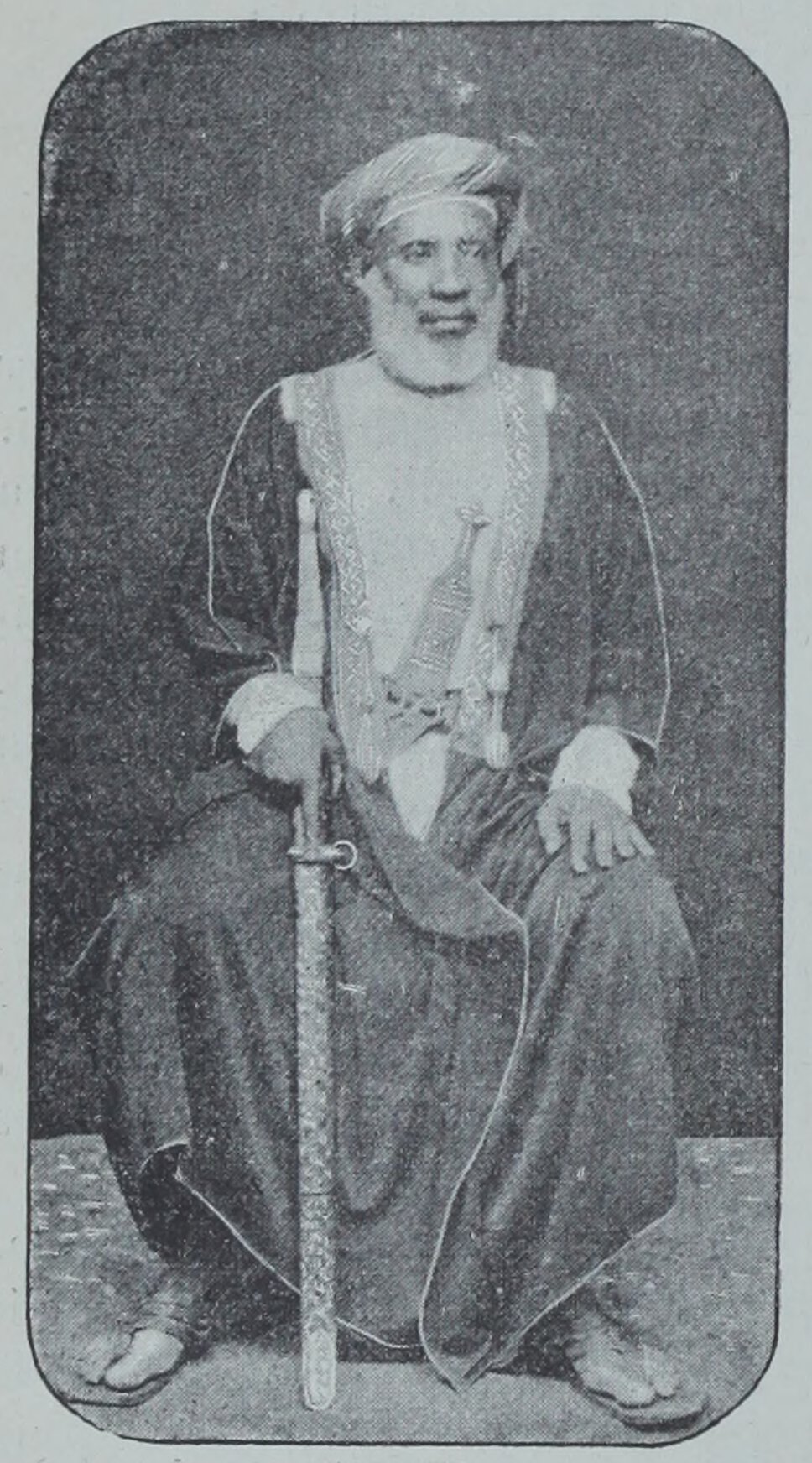 Tippu Tip, seated, facing forward, head turned to his left, holding long sword that touches ground.