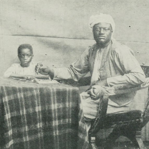 Apolo Kagwa and his son, both seated at a table; Kagwa holds a pen above a notebook.