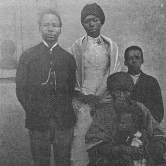 Tausé Soga standing with man and child at her sides and with older woman seated with child on lap.