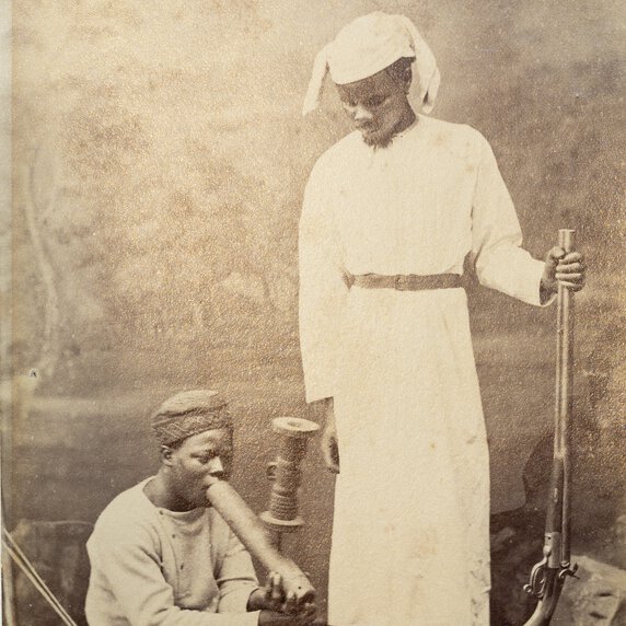 Chuma, seated with legs extended, smoking pipe and being watched by Susi who holds rifle upright.