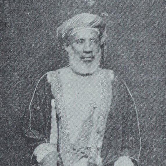 Tippu Tip, seated, facing forward, head turned to his left, holding long sword that touches ground.